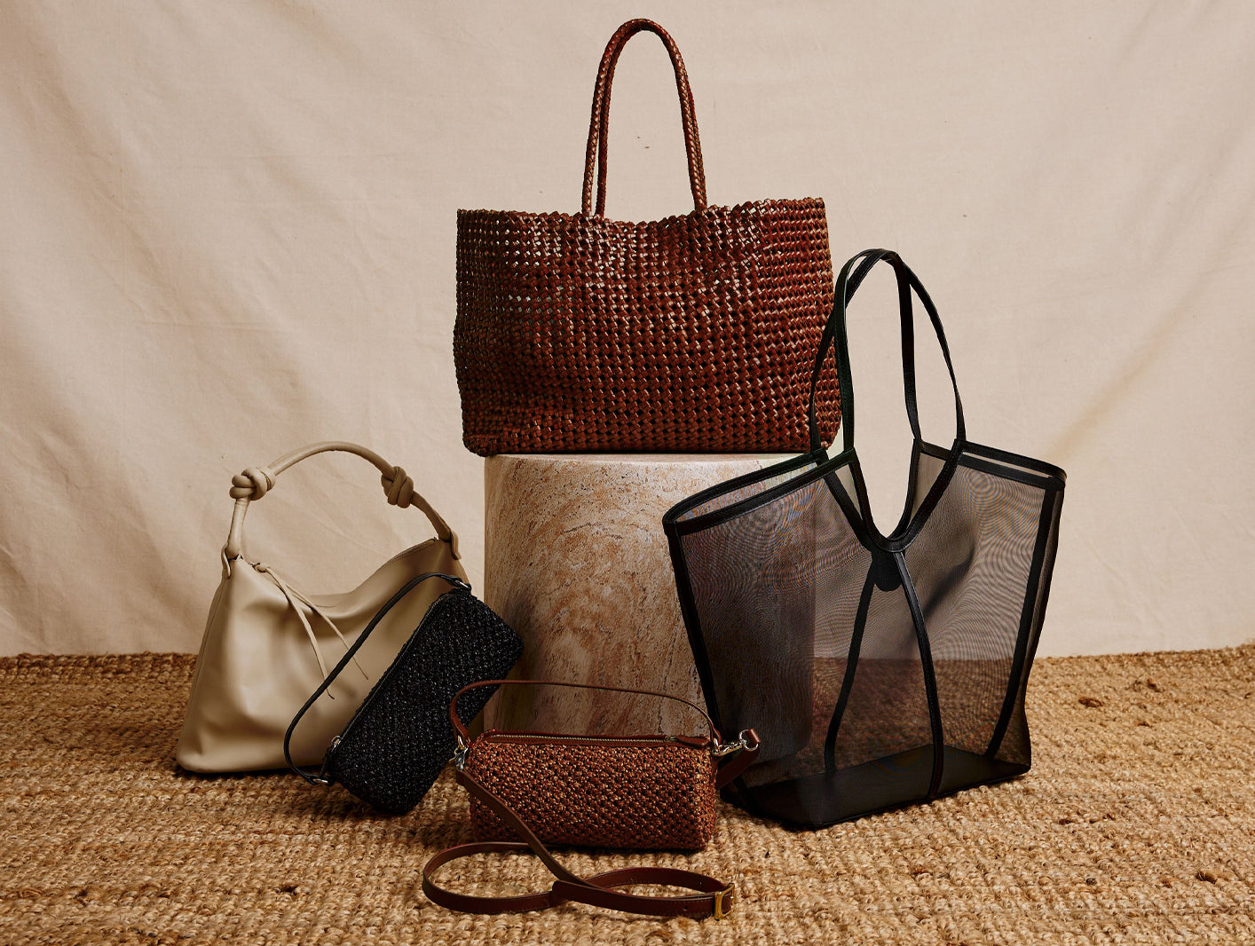Collection of handbags including a woven brown bag, a black mesh tote, a beige bucket bag, and a small brown purse on a textured surface.