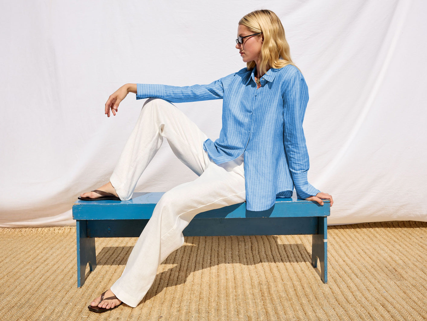 Woman sitting on a blue bench wearing a light blue shirt and white pants against a white background.