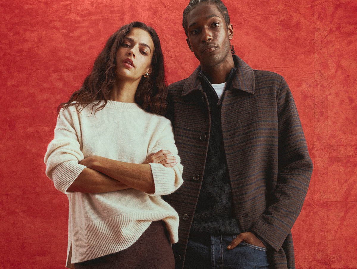 A man and a woman posing together against a textured red-orange background. The woman wears a cream-colored knit sweater, and the man wears a dark brown plaid button-up overshirt over a dark shirt and jeans.