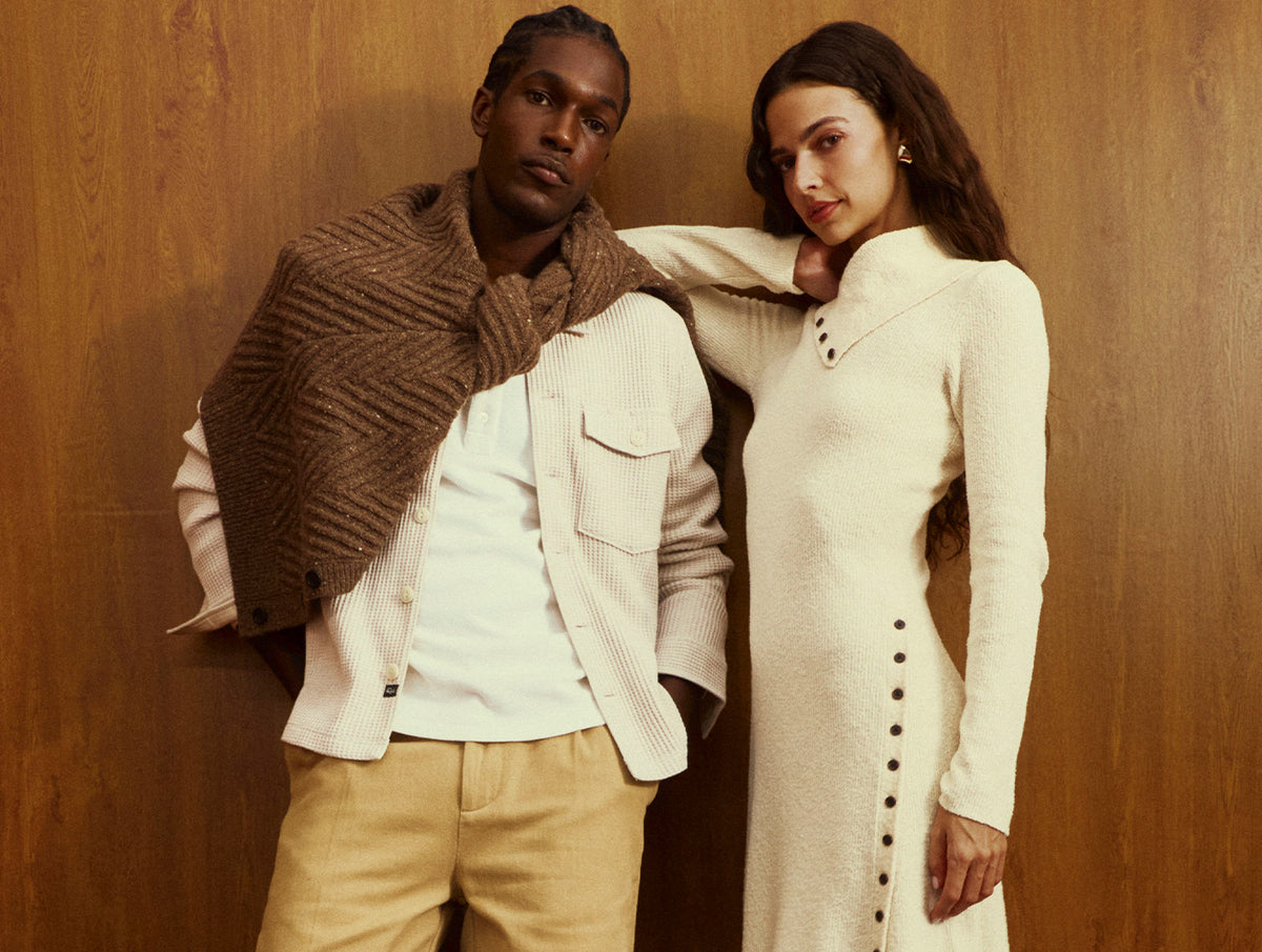 A man and a woman posing together against a warm wood-paneled wall. The man wears a cream ribbed jacket over a white shirt with a brown herringbone knit shawl draped over his shoulders, paired with beige trousers.