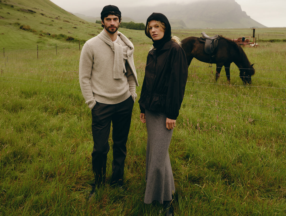 Two models standing in a grassy field with a horse and mountains in the background, wearing dark and light-colored knit outerwear and trousers.