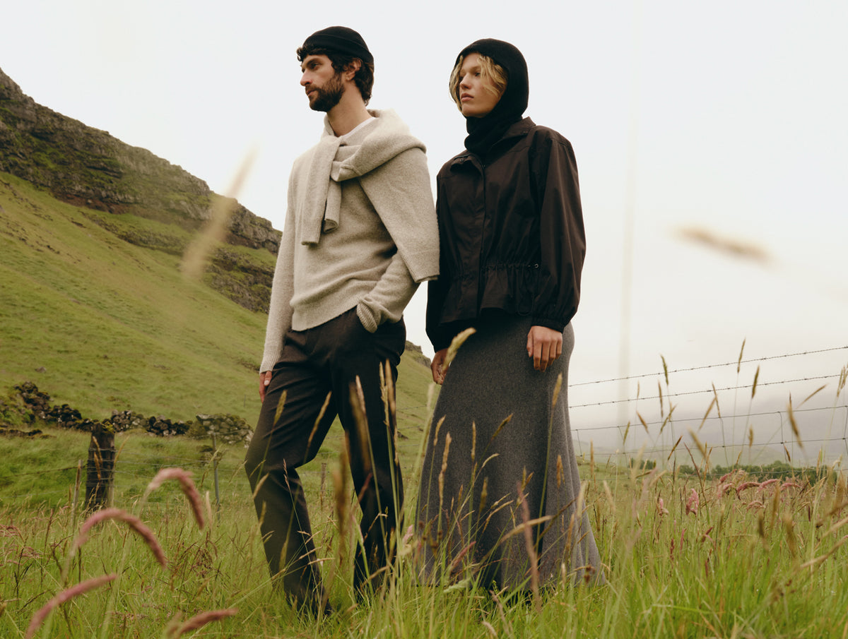 Two models standing in a grassy field with a hillside in the background, wearing earthy-toned outerwear. One wears a light beige knit sweater and dark trousers, and the other wears a dark brown hooded jacket and a long dark skirt.