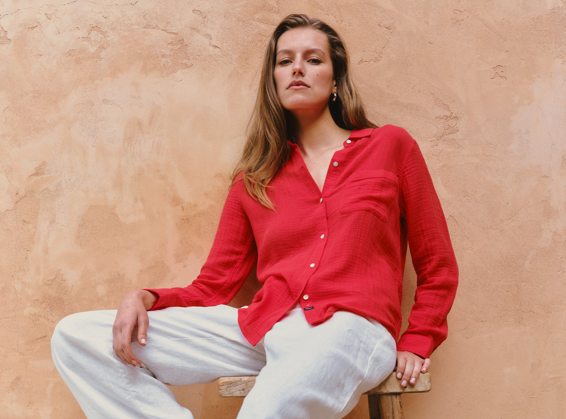 Woman wearing a red shirt and white pants sitting against a beige wall.