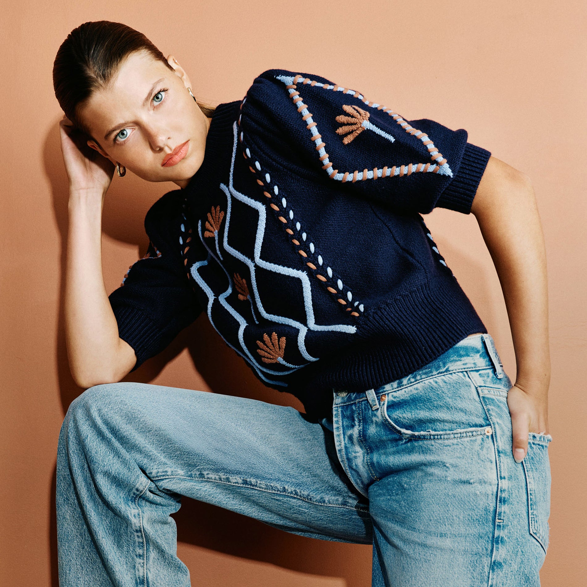 A model wearing a navy blue short-sleeve knit sweater with geometric and floral patterns, paired with light-wash blue jeans, posed against a solid warm peach background.