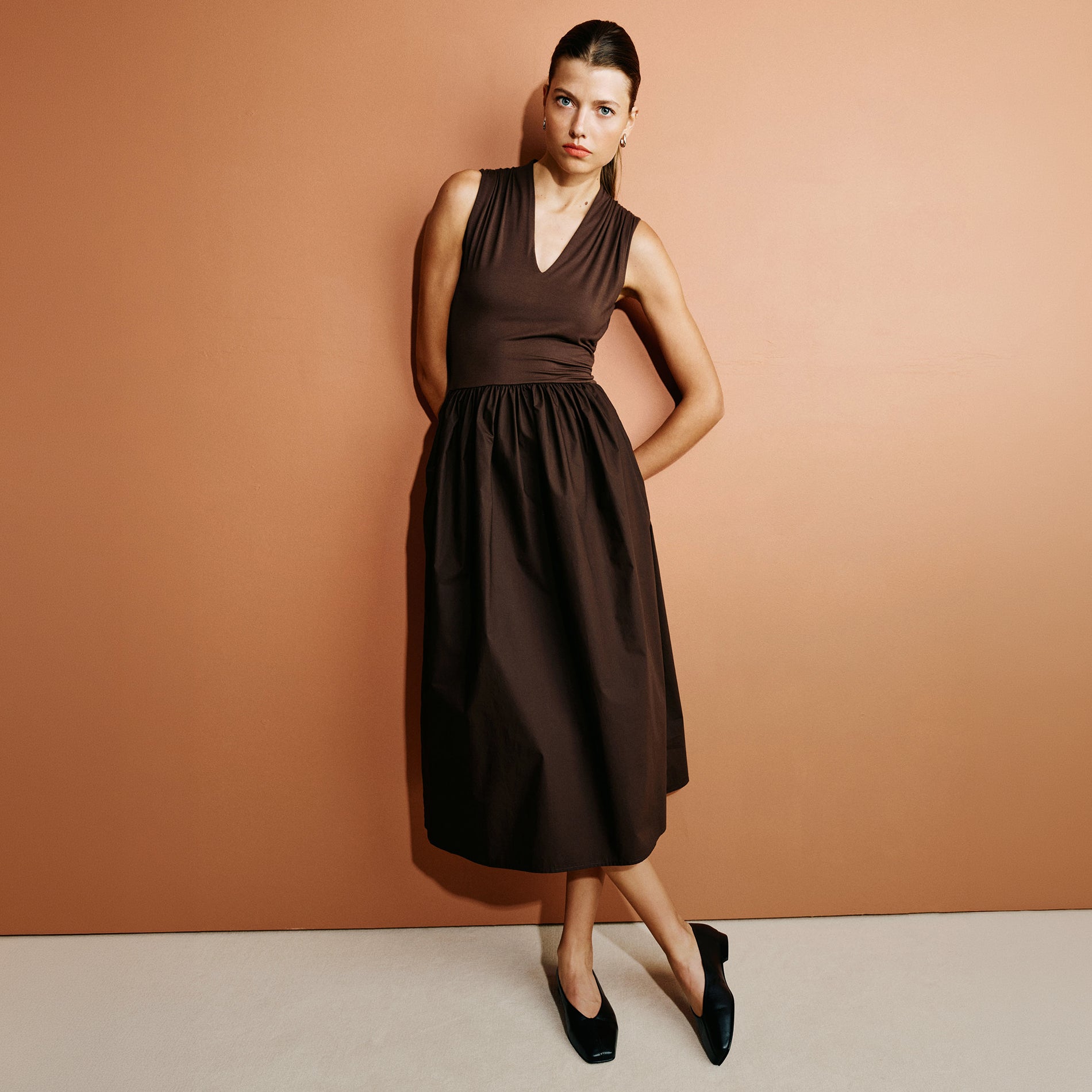 A woman models a sleeveless brown midi dress with a V-neckline and gathered waist, standing against a warm terracotta background.