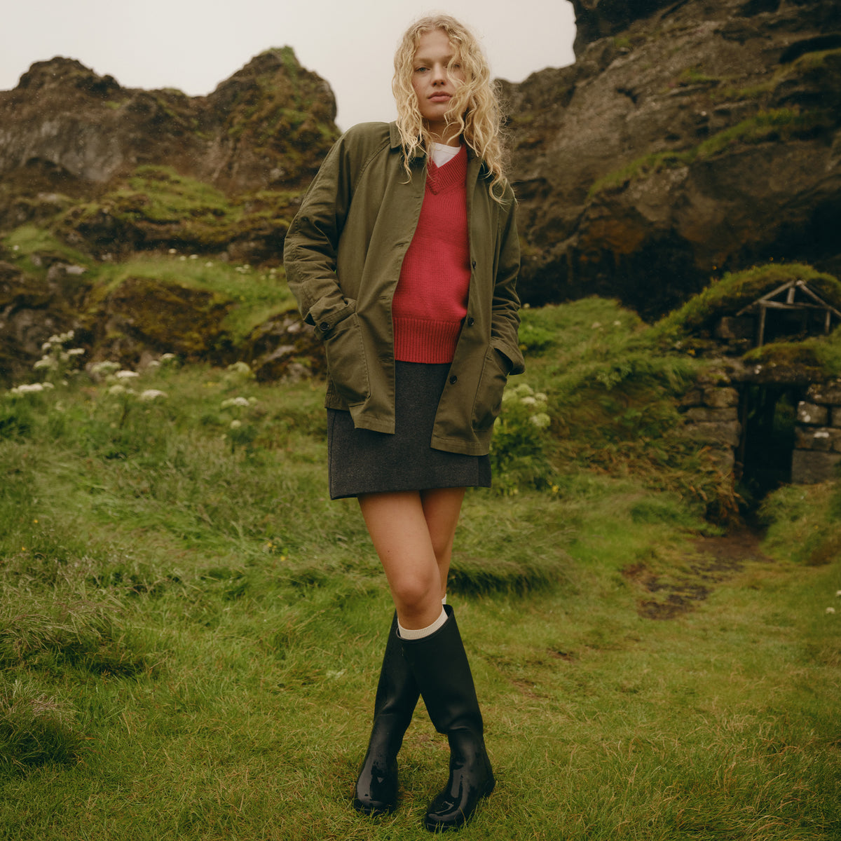 A woman stands outdoors in a grassy, rocky landscape wearing an olive green jacket over a red sweater and dark skirt, with black rain boots.