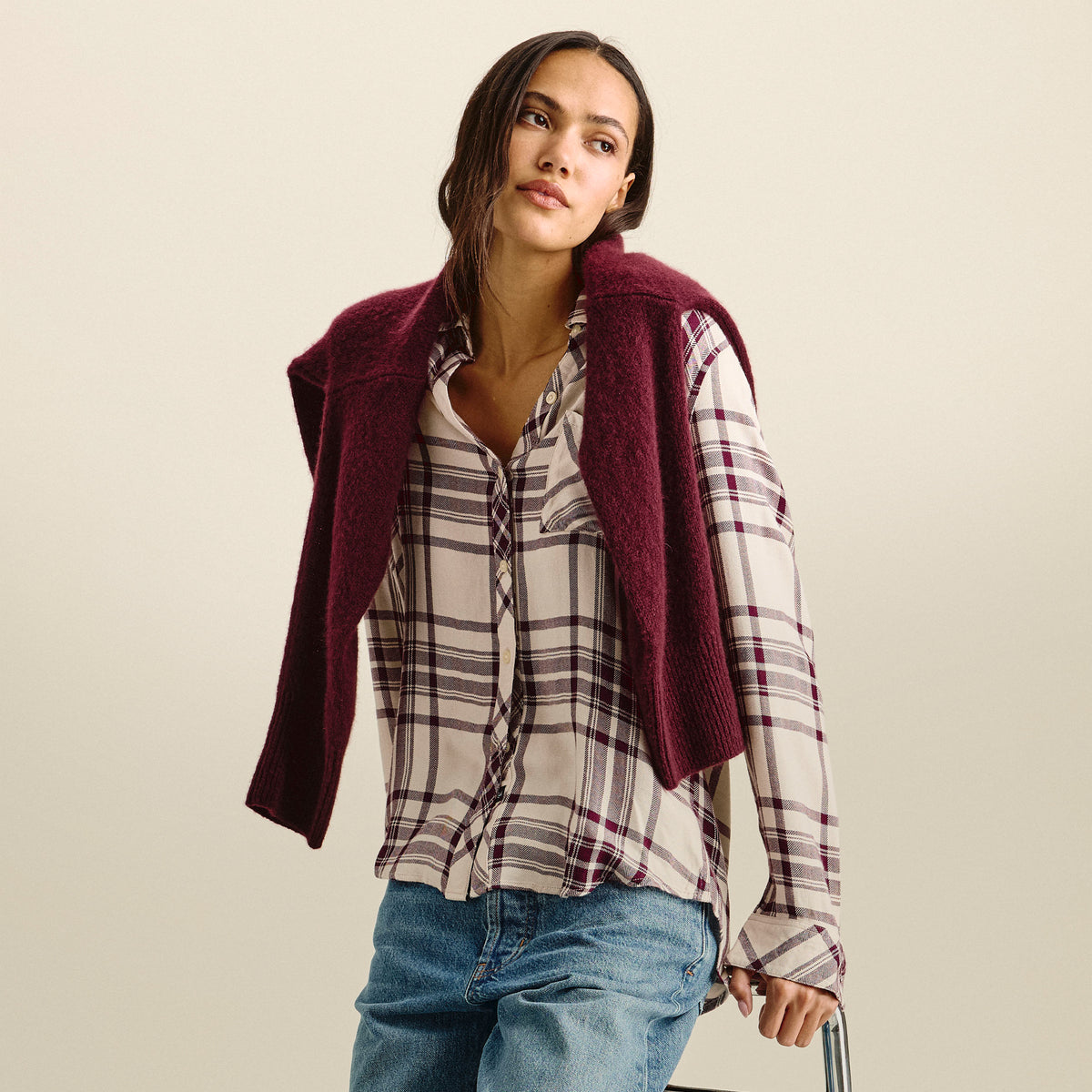 A woman wearing a burgundy fuzzy knit shrug vest over a beige plaid button-down shirt and blue jeans, standing against a solid light beige background.