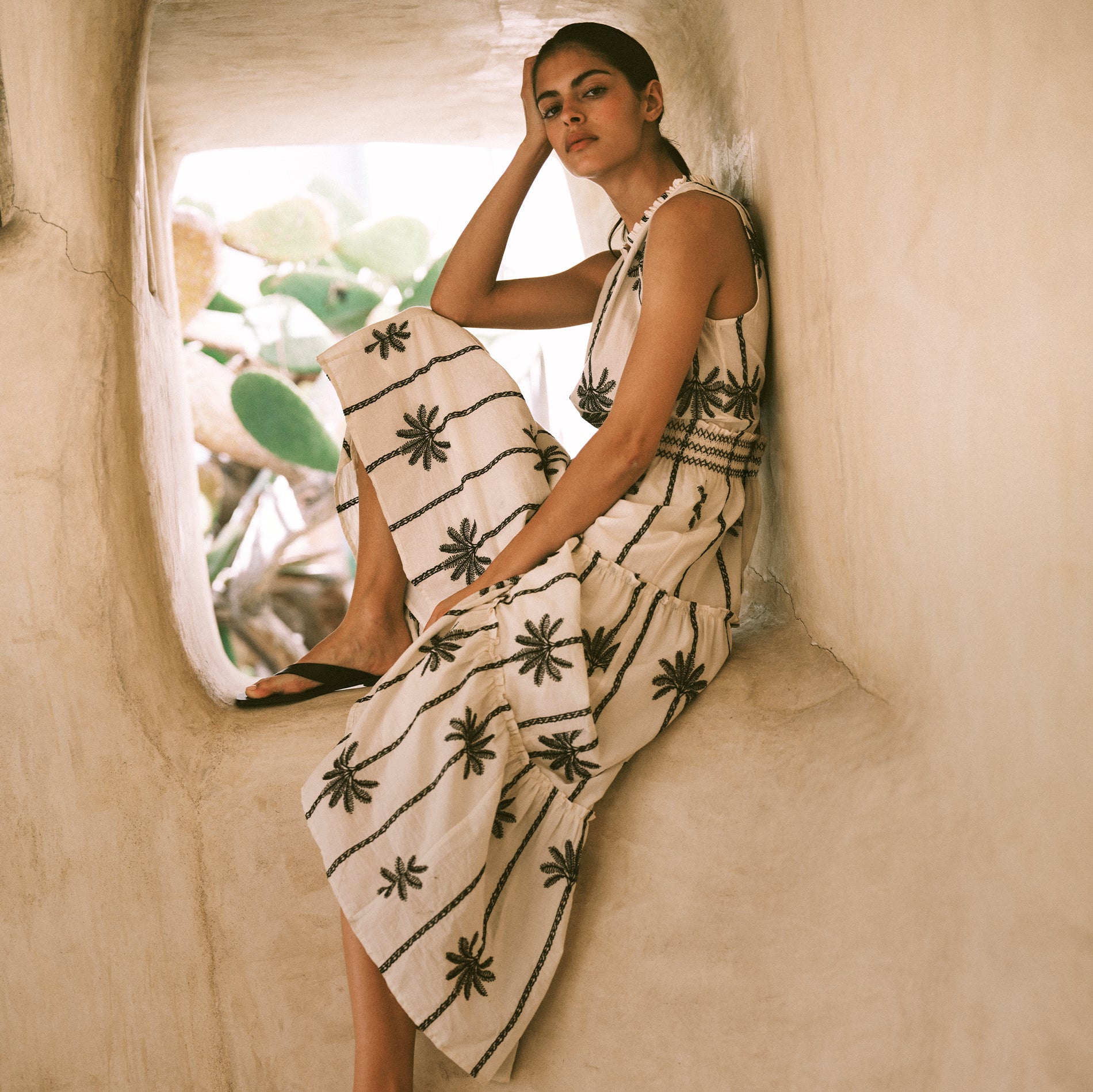 A woman wearing a sleeveless cream maxi dress with black palm tree prints sits in a rustic beige stone alcove.