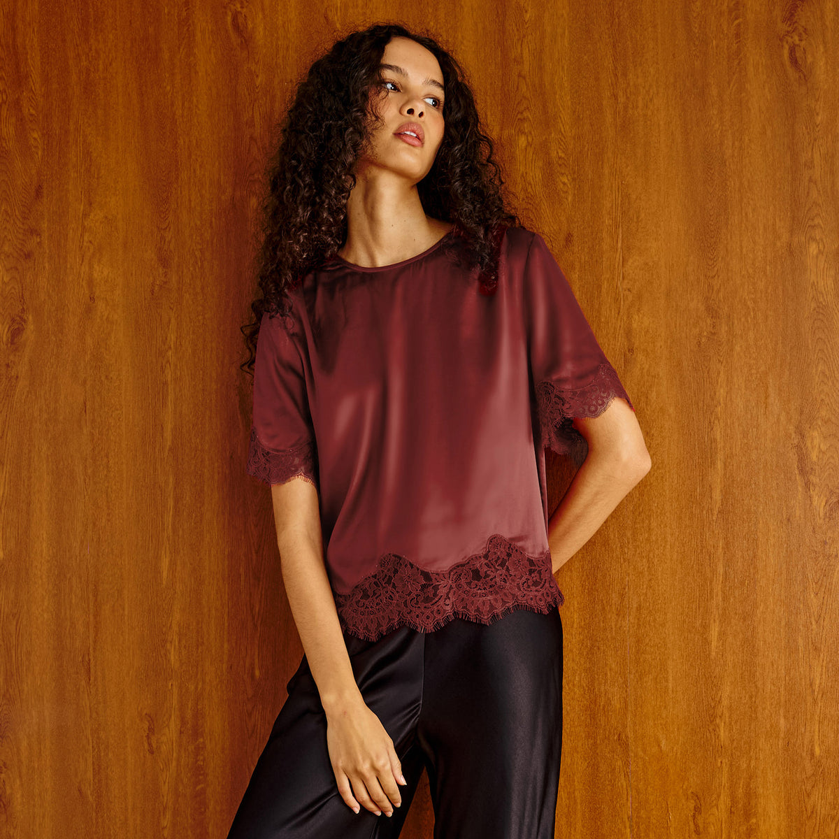 A woman with curly dark hair models a short-sleeve burgundy satin top with lace trim at the hem and cuffs, paired with black satin pants, set against a warm wood-paneled background.