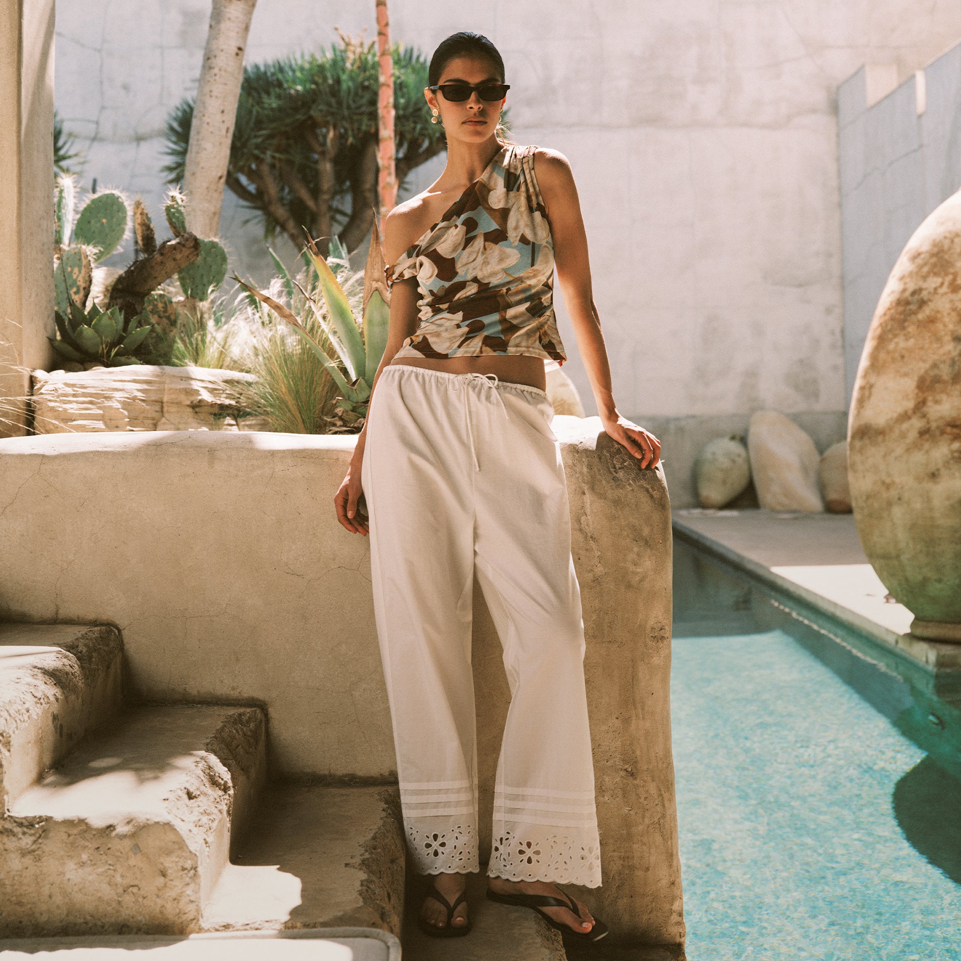 A woman wearing a sleeveless floral print halter top and white wide-leg drawstring pants stands beside a swimming pool in a sunlit outdoor setting with stone walls and plants.