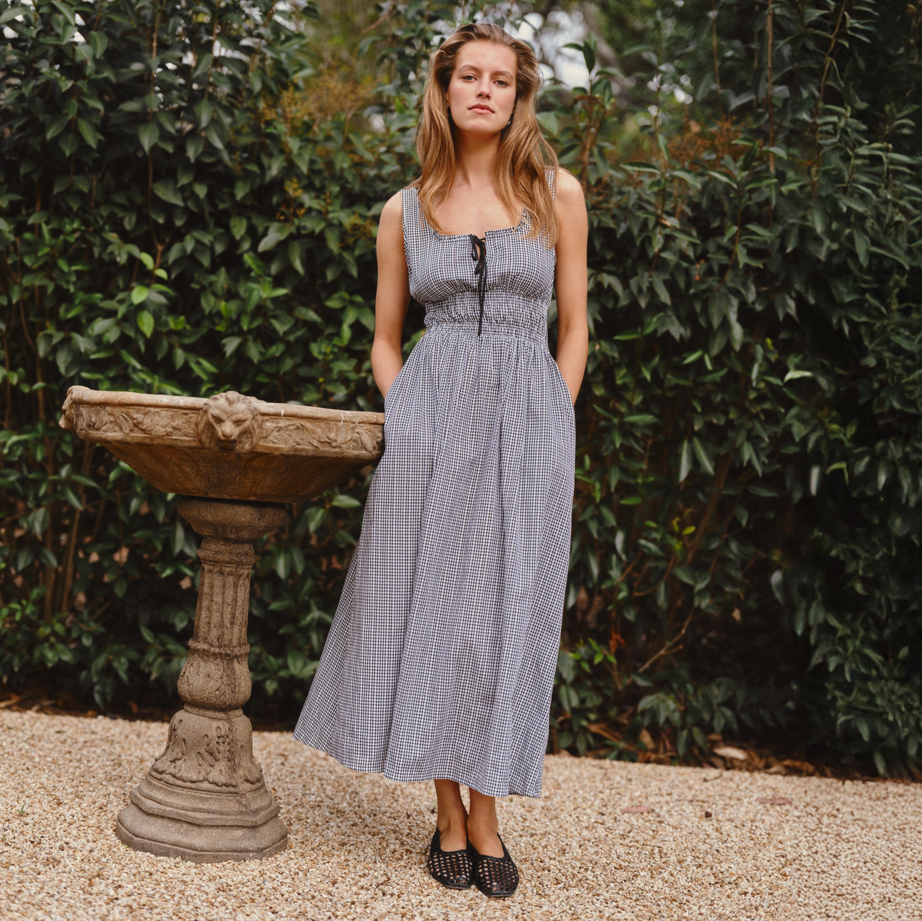 Woman in a checkered dress standing in a garden with a stone fountain.