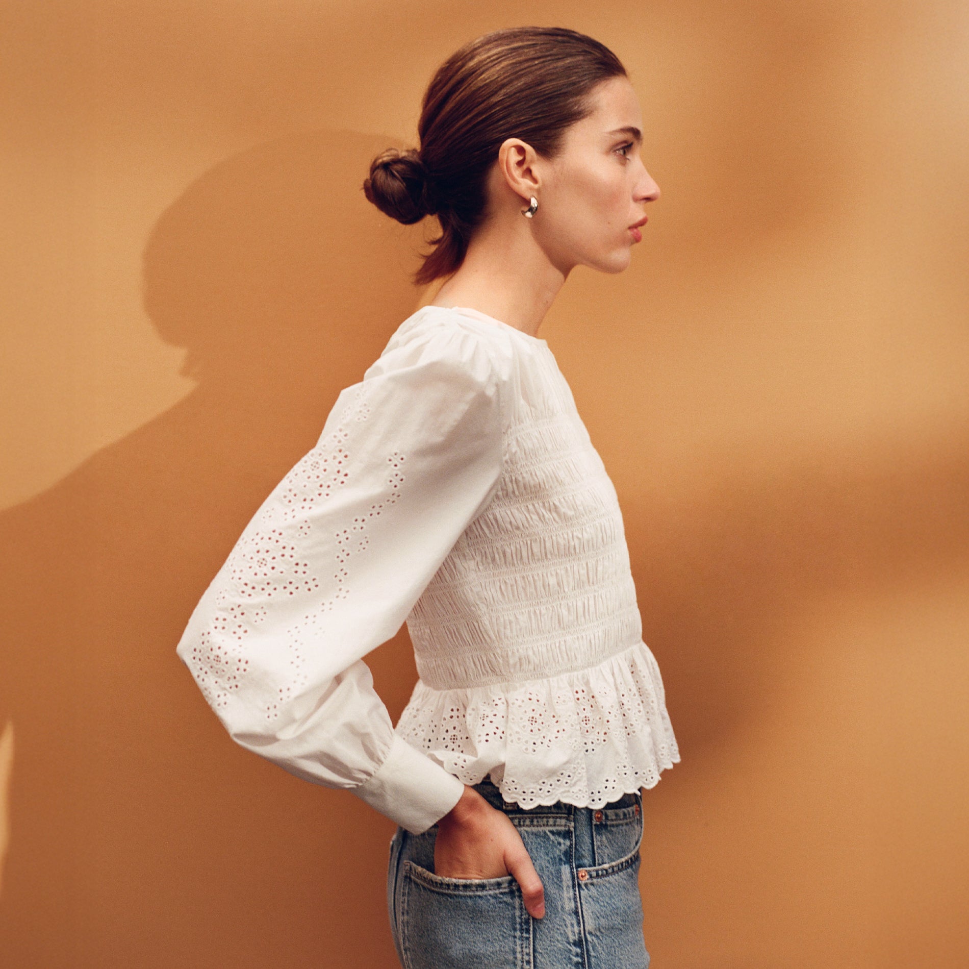 Woman wearing a white blouse with long sleeves and a ruffled bottom against a brown background.