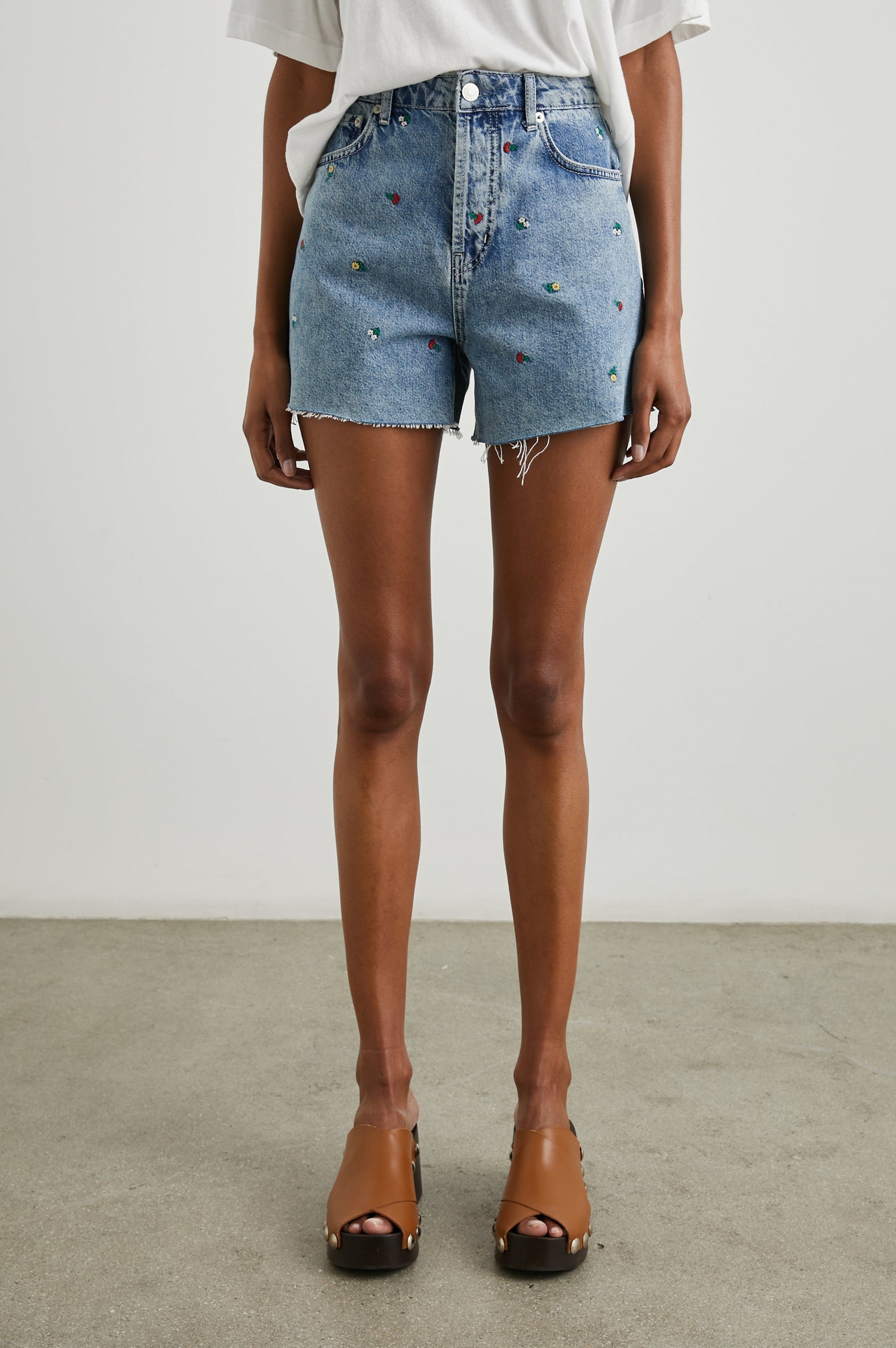 Zuma Denim Shorts with a Raw Hem, Belt Loops, Two Front Pockets Plus a Coin Pocket and Two Back Patch Pockets in Flower Field Colorway, with Embroidered Yellow, White and Red Daisies - Front View Featured on Model 