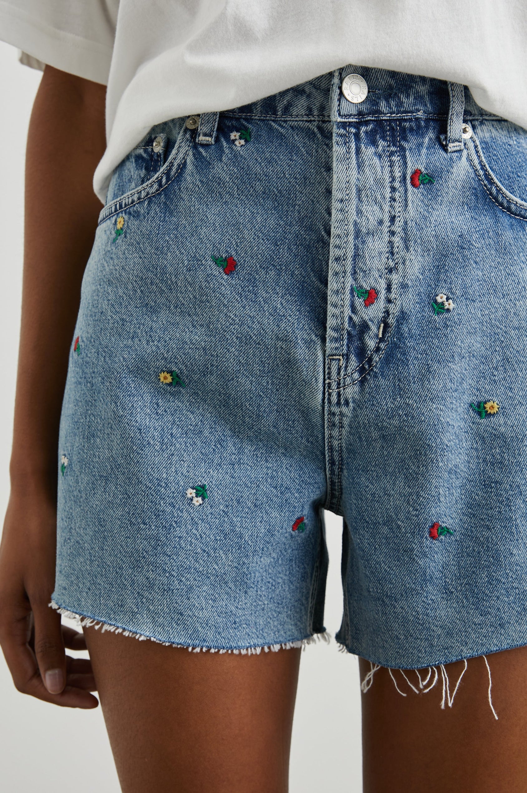 Zuma Denim Shorts with a Raw Hem, Belt Loops, Two Front Pockets Plus a Coin Pocket and Two Back Patch Pockets in Flower Field Colorway, with Embroidered Yellow, White and Red Daisies - Front View Featured on Model 