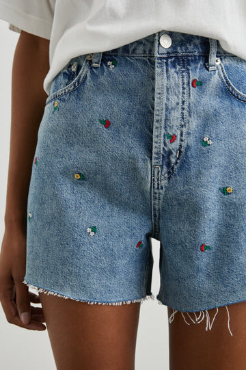 Zuma Denim Shorts with a Raw Hem, Belt Loops, Two Front Pockets Plus a Coin Pocket and Two Back Patch Pockets in Flower Field Colorway, with Embroidered Yellow, White and Red Daisies - Front View Featured on Model 