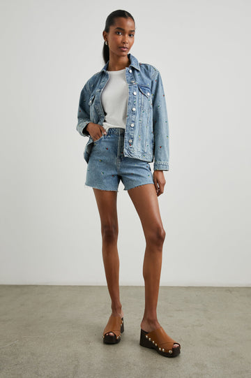 Zuma Denim Shorts with a Raw Hem, Belt Loops, Two Front Pockets Plus a Coin Pocket and Two Back Patch Pockets in Flower Field Colorway, with Embroidered Yellow, White and Red Daisies - Front Full Body View Featured on Model 