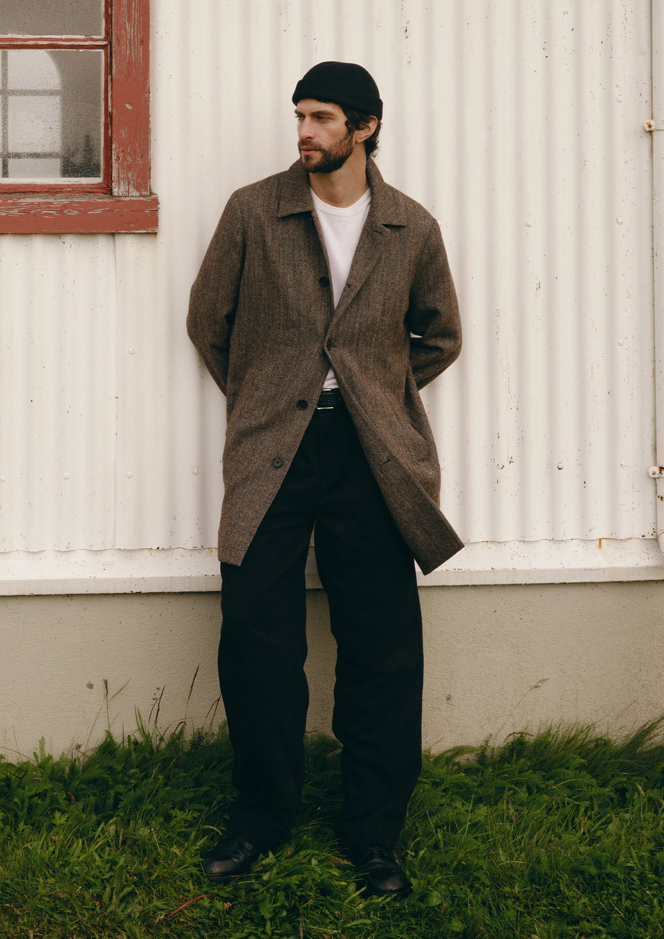 A man wearing a brown textured overcoat, black trousers, a white t-shirt, and a black beanie stands against a white corrugated metal wall with green grass at the bottom.
