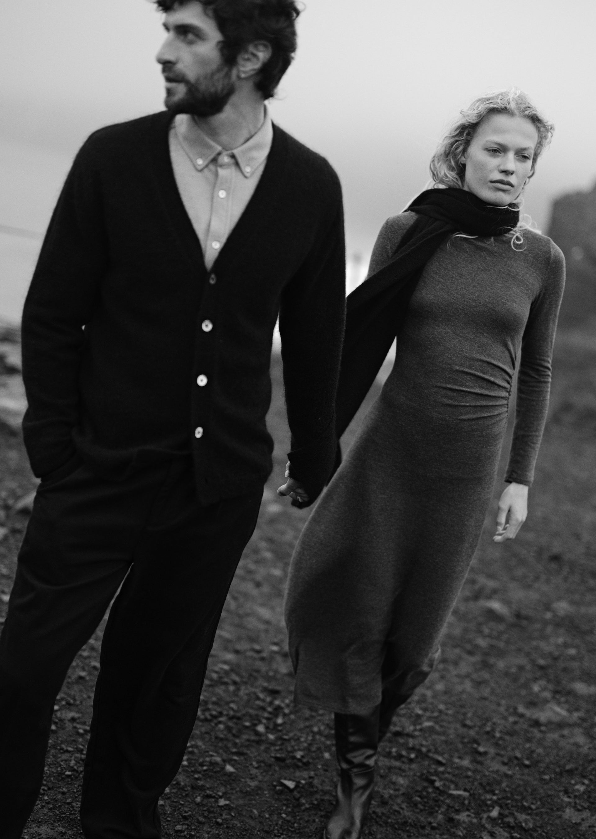A black and white lifestyle photograph of a man and a woman walking hand-in-hand outdoors. The man wears a dark cardigan over a collared shirt and trousers, while the woman wears a dark long-sleeve dress and scarf.