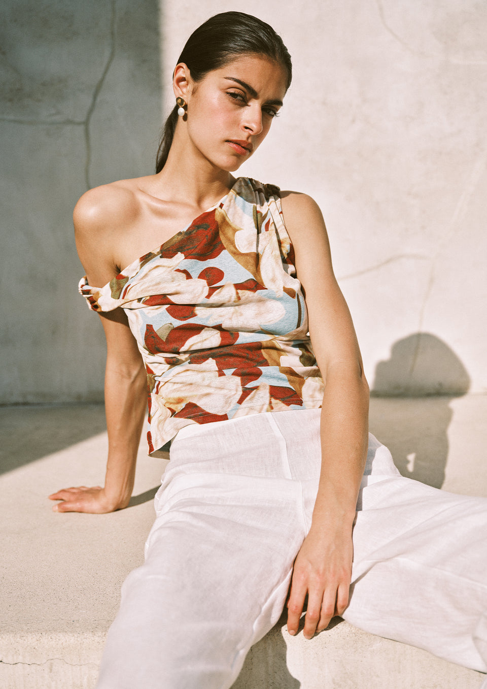 Woman wearing a patterned top and white pants sitting on a stone surface.