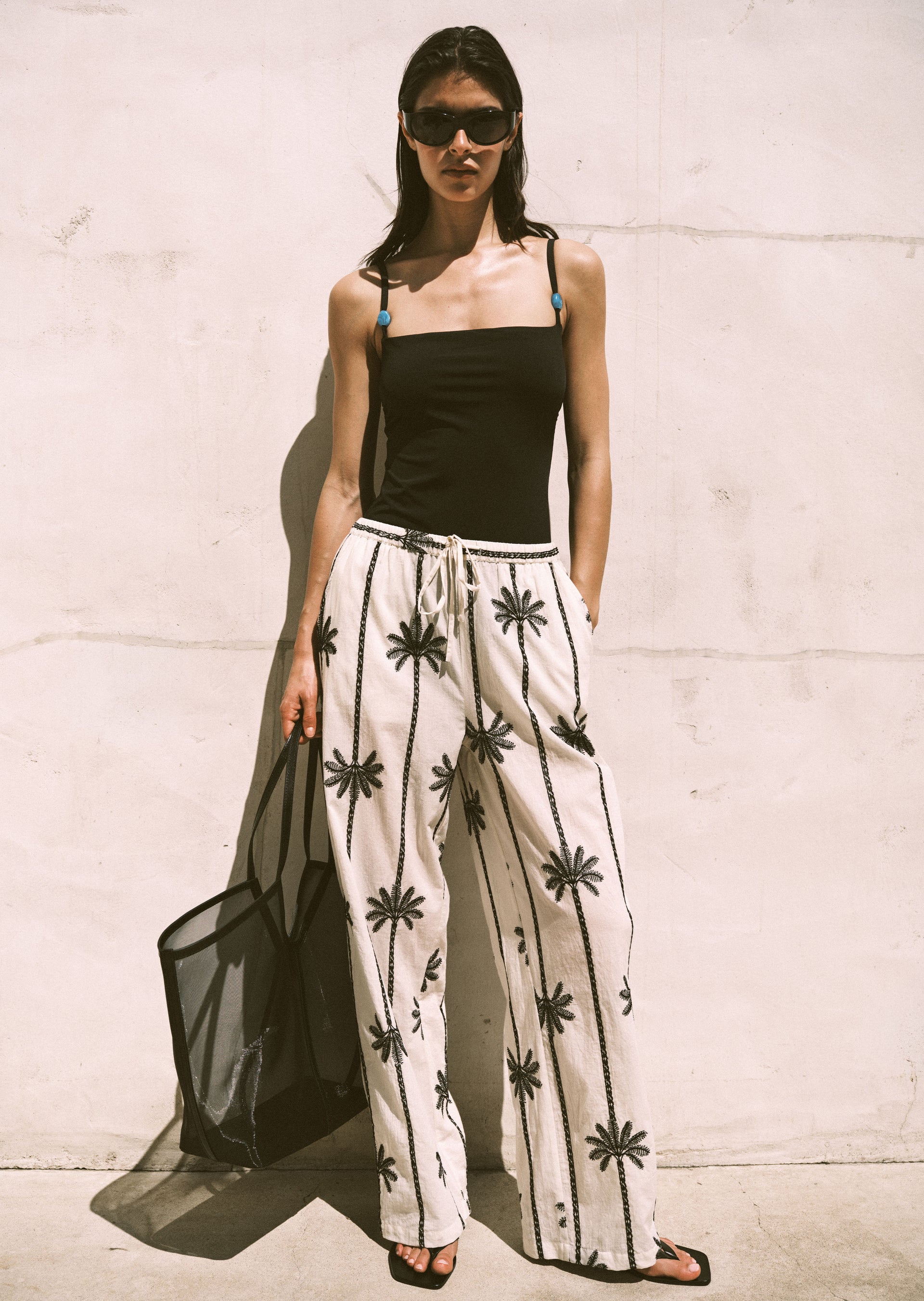 Woman wearing a black swimsuit and beige pants with black patterns against a light-colored wall.