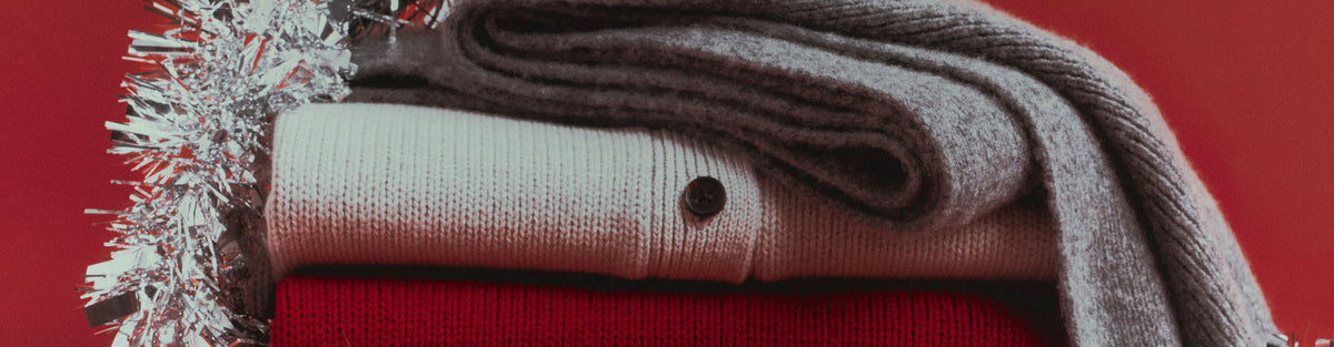 Folded stack of knits in cream, red, and gray, topped with a black beanie and a gray scarf, wrapped in silver tinsel against a red background.