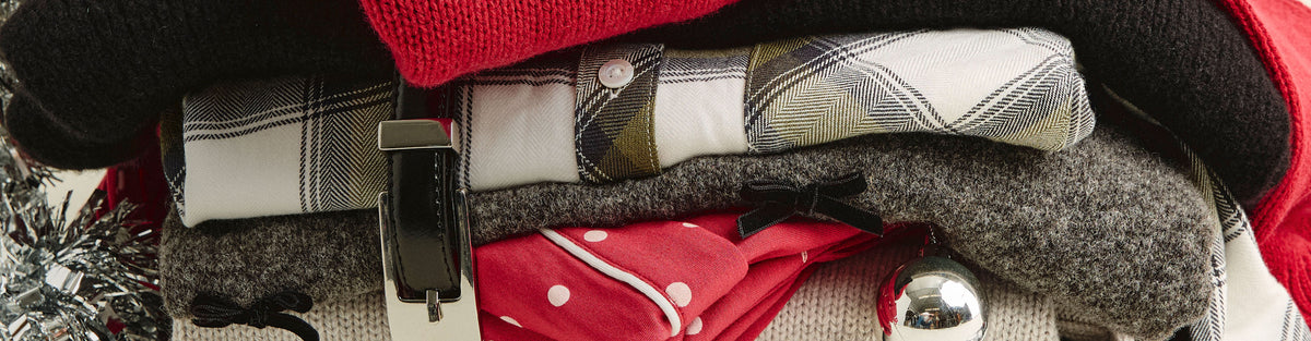 Close-up of a folded stack of holiday clothing, including a red sweater, black knit, plaid shirt, gray sweater with a small black bow, and red polka-dot pajamas, with a black leather belt and silver ornament.