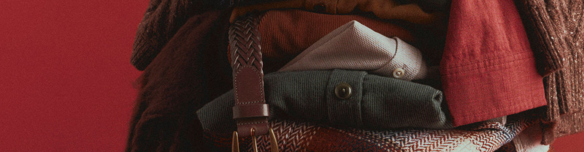Close-up of a folded stack of menswear in olive, rust, cream, and red, with denim, a braided brown leather belt hanging down the front, and a red plaid piece at the bottom, against a red background.