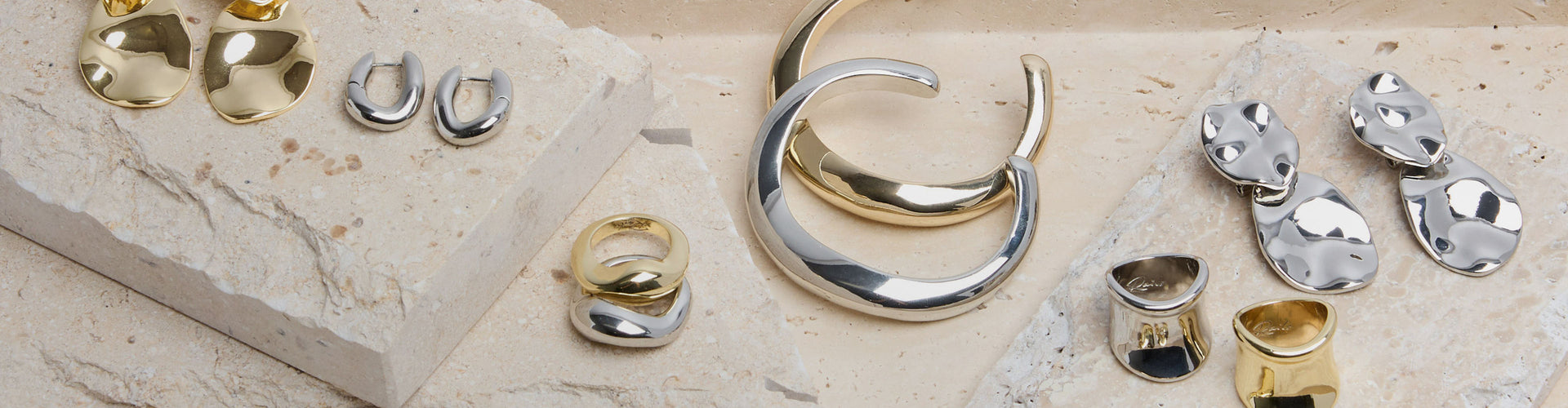 A styled flat-lay of various gold and silver jewelry pieces, including hoop earrings, rings, and geometric stud earrings, arranged on textured beige stone slabs.