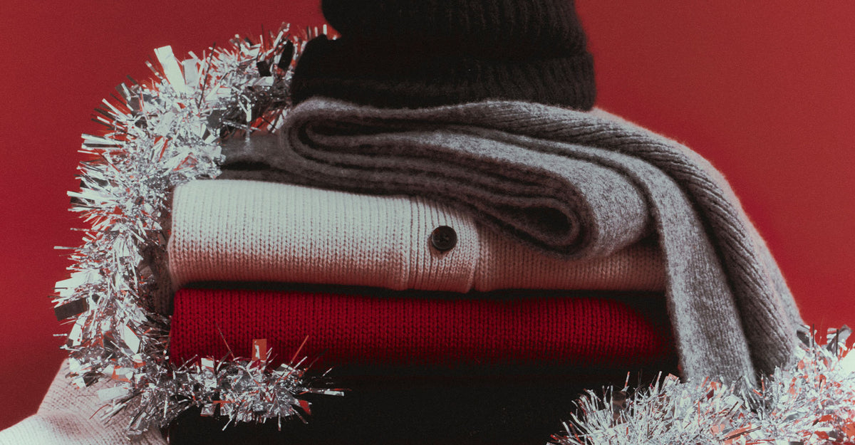 Folded stack of knits in cream, red, and gray, topped with a black beanie and a gray scarf, wrapped in silver tinsel against a red background.