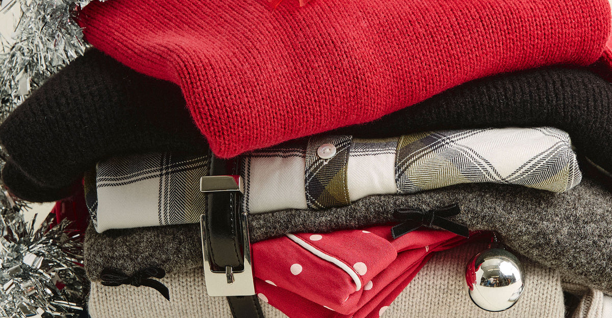 Close-up of a folded stack of holiday clothing, including a red sweater, black knit, plaid shirt, gray sweater with a small black bow, and red polka-dot pajamas, with a black leather belt and silver ornament.