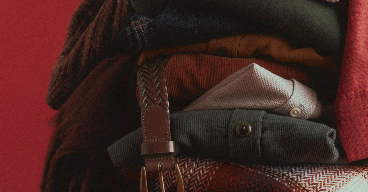 Close-up of a folded stack of menswear in olive, rust, cream, and red, with denim, a braided brown leather belt hanging down the front, and a red plaid piece at the bottom, against a red background.