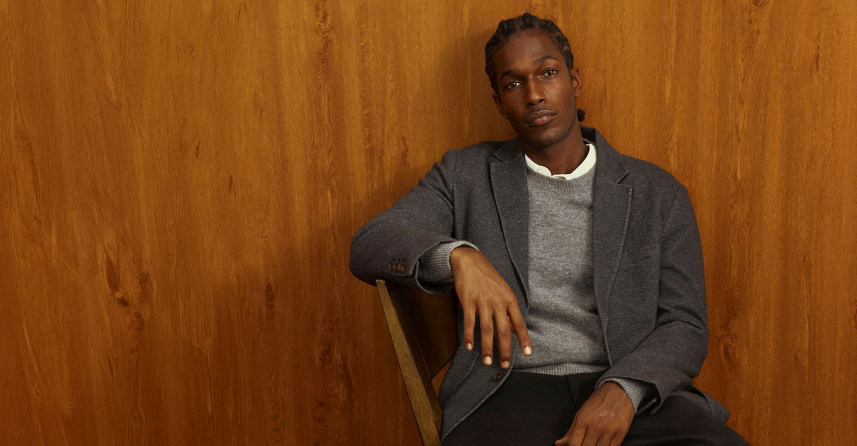 Man seated on a wooden chair in a gray blazer over a gray crewneck sweater and white collared shirt, against a warm wood-paneled wall.