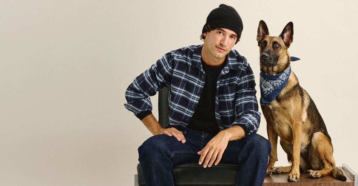 Seated man in a navy and light blue plaid flannel shirt, black tee, black beanie, and dark denim, with a German Shepherd in a navy bandana beside him on a cream backdrop.
