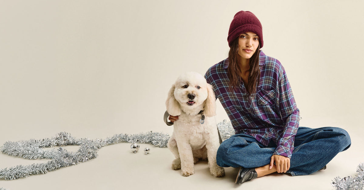 Woman sitting cross-legged in a purple plaid shirt, burgundy beanie, wide-leg jeans, and black loafers, with a fluffy white dog beside her, surrounded by silver tinsel and ornaments on a cream backdrop.
