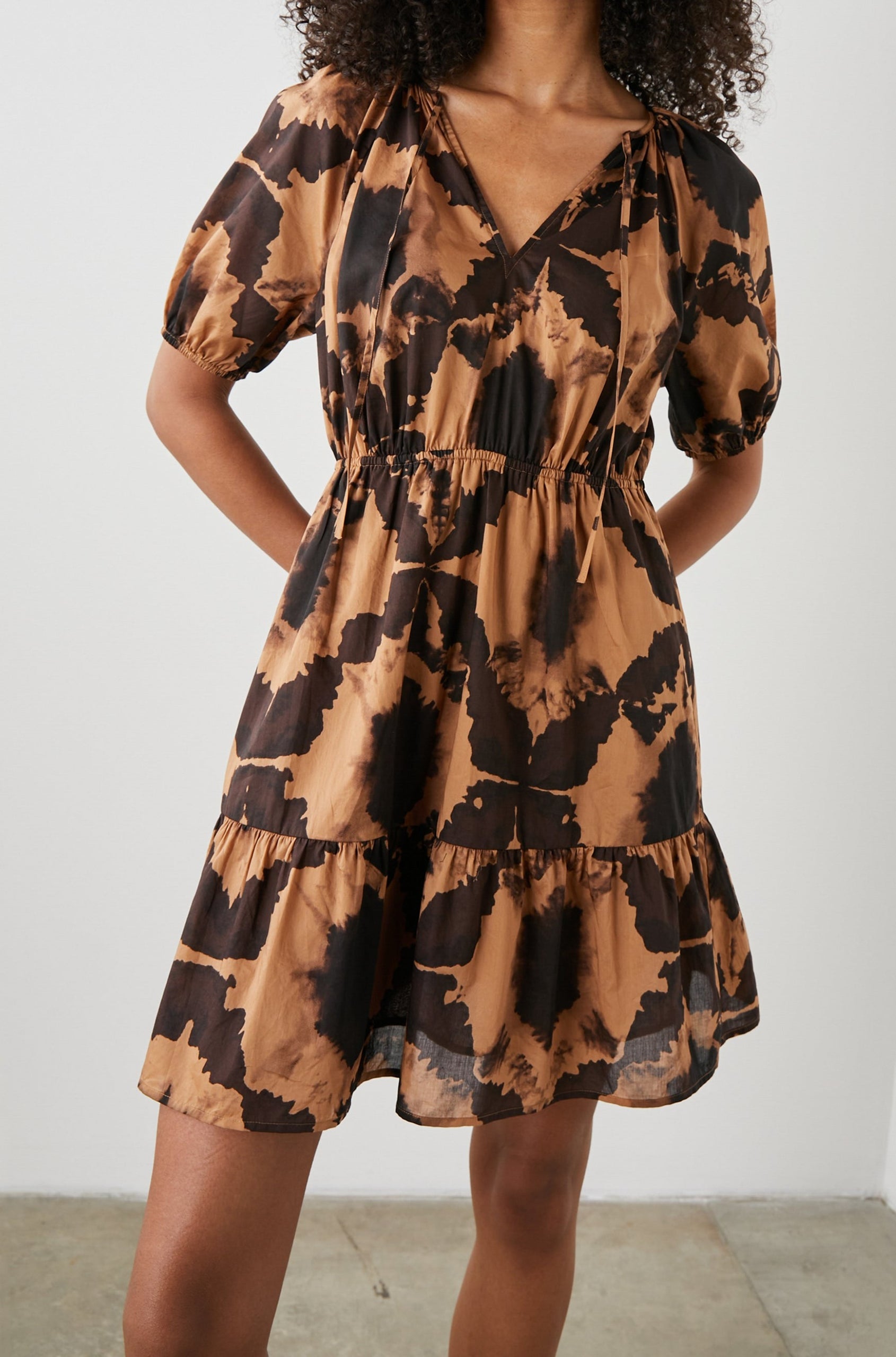 Amabella Short Sleeve Mini Dress with a V neck with Ties, Puff Sleeves, a Cinched Waist, and a Tiered Skirt in Tigers Eye Colorway, a Floral Pattern in Browns on Dark Brown - Front View Featured on Model  
