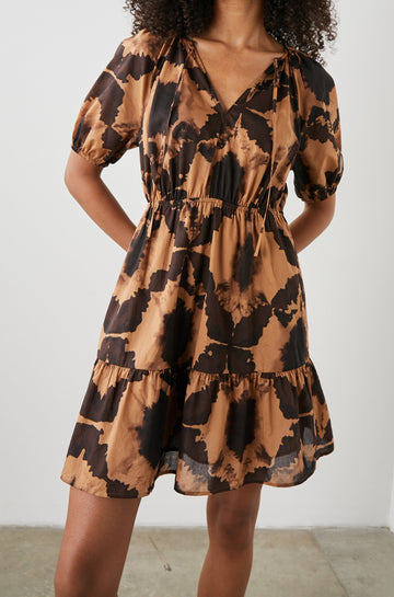 Amabella Short Sleeve Mini Dress with a V neck with Ties, Puff Sleeves, a Cinched Waist, and a Tiered Skirt in Tigers Eye Colorway, a Floral Pattern in Browns on Dark Brown - Front View Featured on Model  