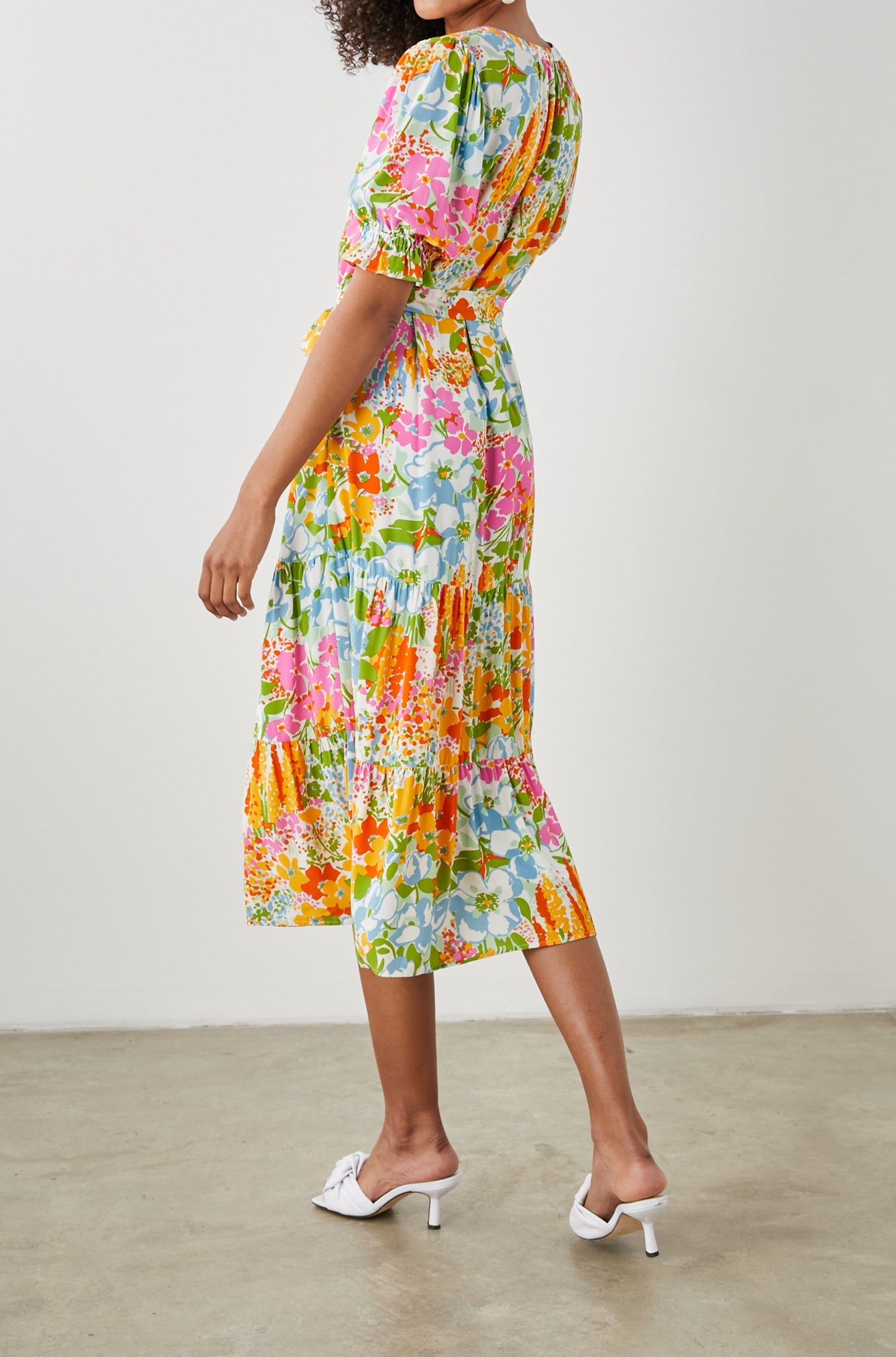 Eliana Midi Dress with Elbow-Length Sleeves with Shirred Cuffs, a V Neck, Buttons Down the Bodice, a Fabric Tie at the Waist  and a Tiered Skirt in Day Garden Colorway, a Floral Pattern in Blue, Pink and Oranges on White - Back Full Body View Featured on Model